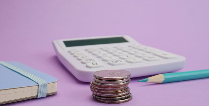 Cálculos financeiros, moedas empilhadas, calculadora, caderno e lápis em fundo roxo para serviços de gestão tributária e contábil da Dootax.